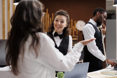 Elderly lady tourist checks in at hotel lobby registration desk, anxious to begin delightful vacation. Smiling young concierge providing advice to retired senior woman for best holiday activities.